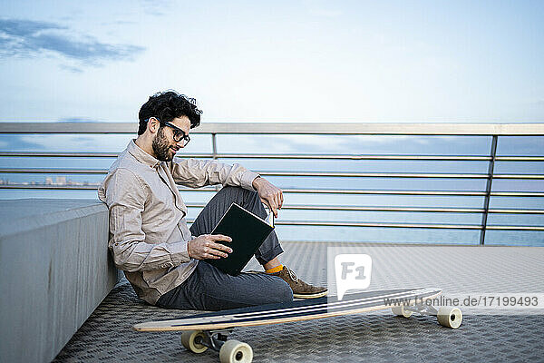 Geschäftsmann  der Tagebuch liest  während er mit seinem Longboard am Pier sitzt