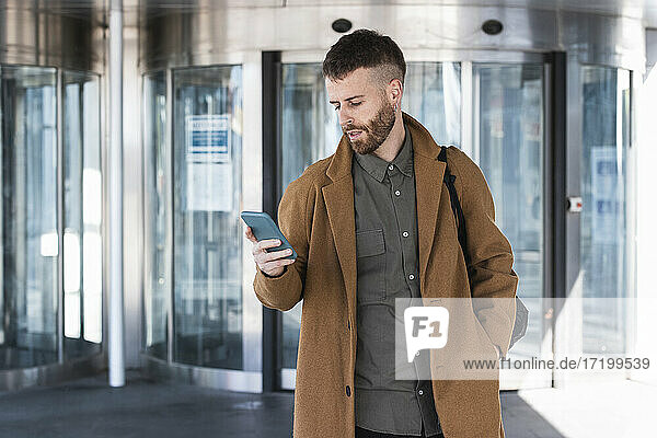 Geschäftsmann mit Jacke  der ein Mobiltelefon benutzt  während er mit den Händen in den Taschen vor einem Gebäude steht
