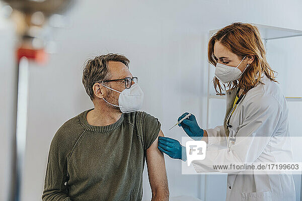 Allgemeinmediziner injiziert COVID-19-Impfstoff in den Arm eines Patienten  während er im Untersuchungsraum steht