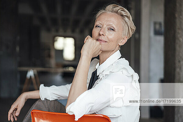 Blond businesswoman with hand on chin sitting on chair on loft apartment