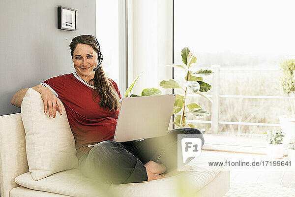 Smiling woman with laptop wearing headphones while sitting on sofa in living room
