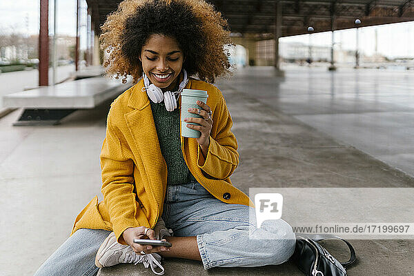 Smiling Afro woman with disposable coffee cup using mobile phone in sitting area