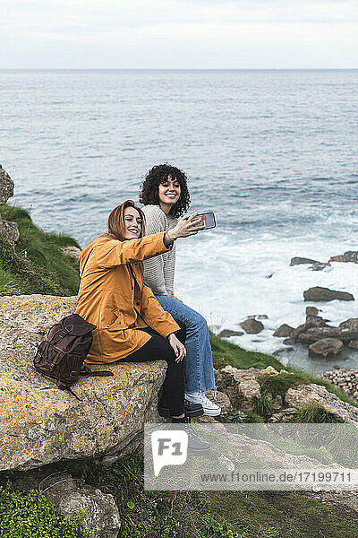 Fröhliche Freundinnen machen ein Selfie  während sie auf einem Felsen am Meer sitzen