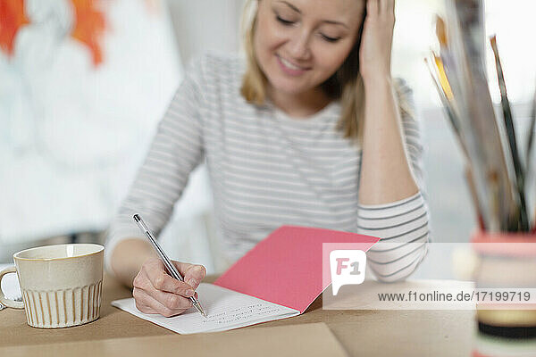 Lächelnde Frau mit Kaffeetasse  die in einem Buch auf dem Tisch schreibt  während sie im Heimstudio sitzt