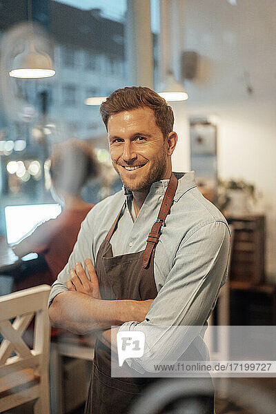 Owner wearing apron smiling while standing with arms crossed at cafe