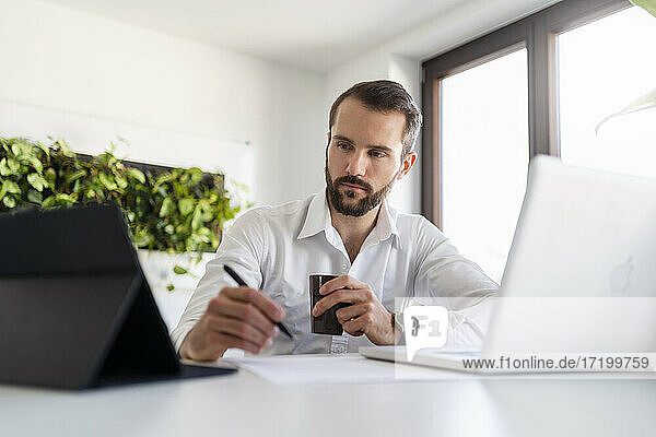 Geschäftsmann mit Laptop und Kaffeetasse bei der Nutzung eines digitalen Tablets im Büro