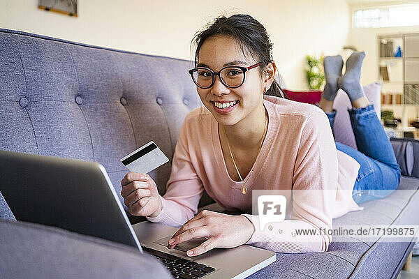 Smiling woman with credit card and laptop relaxing on sofa at home