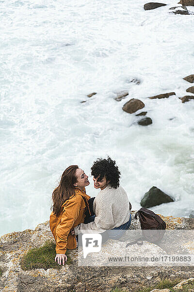 Glückliche junge Freundinnen sitzen auf einem Felsen am Meer während des Wochenendes