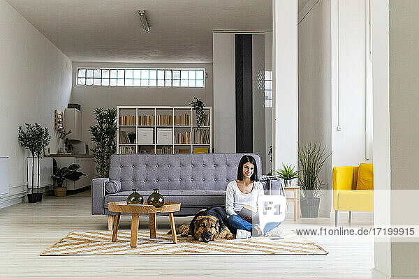 Lächelnde Frau liest ein Buch und sitzt mit ihrem Hund auf dem Teppich im Wohnzimmer