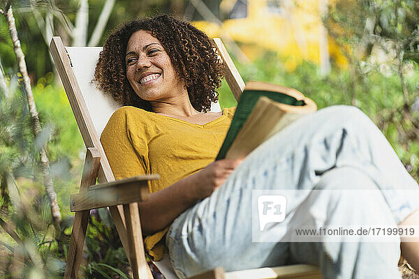 Lächelnde Frau mit lockigem Haar und Buch auf einem Stuhl im Garten sitzend
