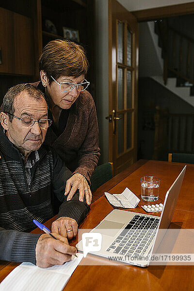 Senior man with social worker writing on book while using laptop at home during