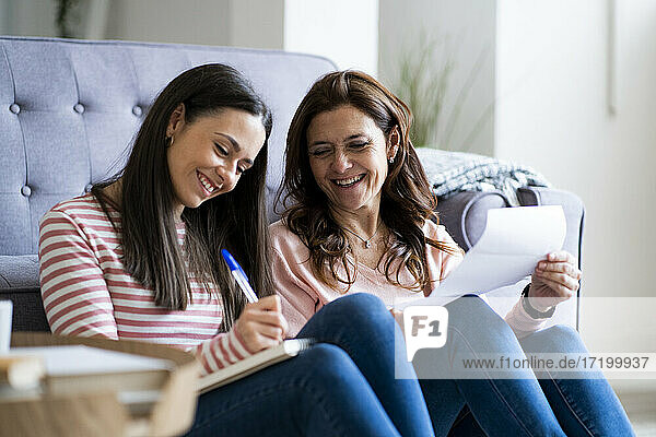 Smiling mother and daughter checking bills while sitting by sofa in living room