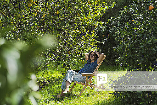 Frau mit Händen hinter dem Kopf entspannt auf einem Stuhl inmitten von Bäumen im Garten