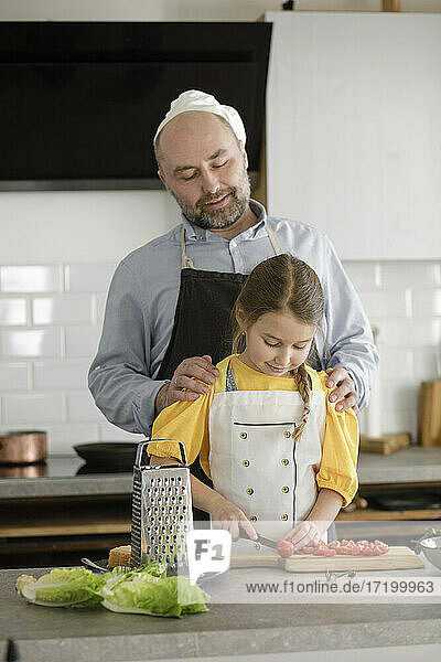 Vater sieht seine Tochter beim Schneiden von Tomaten an  während er in der Küche zu Hause steht