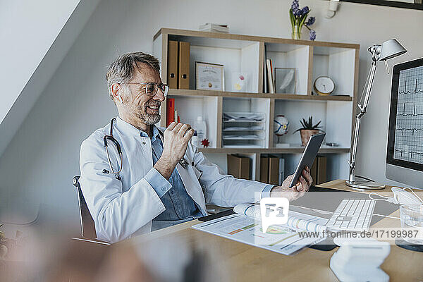 Älterer Arzt  der über ein digitales Tablet einen Videogespräch führt  während er in der Arztpraxis sitzt