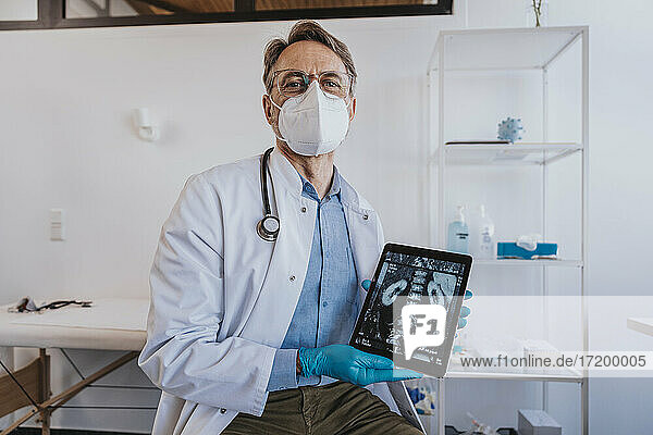 Arzt mit Gesichtsmaske zeigt Röntgenbild auf digitalem Tablet  während er in der Klinik sitzt