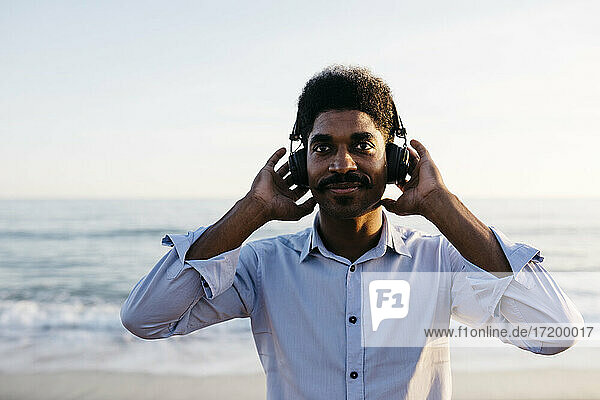Smiling Afro man listening music through headphones at beach against clear sky