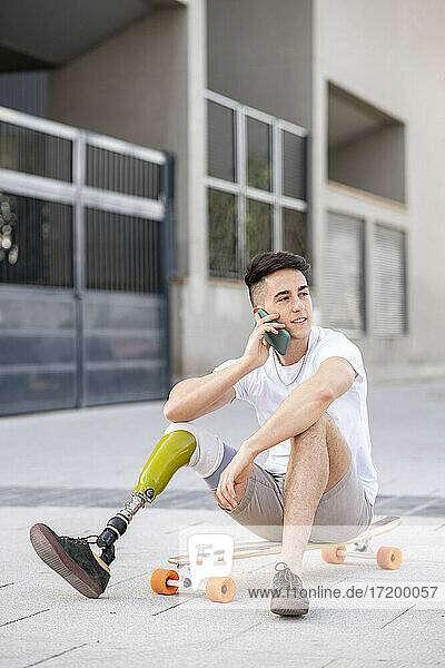 Junger behinderter Mann  der auf einem Skateboard sitzend über sein Handy spricht