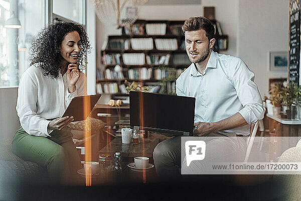Lächelnde Geschäftsfrau mit digitalem Tablet  die neben einem Kollegen sitzt  der in einem Café am Laptop arbeitet