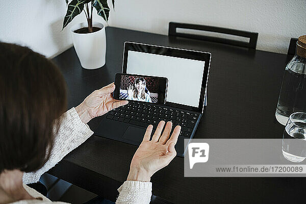 Woman with laptop gesturing while talking with daughter on video call through mobile phone at home