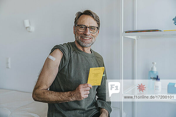 Lächelnder Mann mit Verband am Arm  der den Impfpass zeigt  während er im Untersuchungsraum sitzt