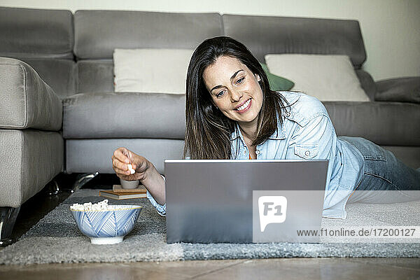 Lächelnde Frau mit Laptop  die Popcorn isst  während sie zu Hause auf dem Teppich liegt