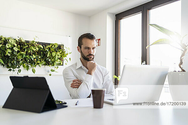 Nachdenklicher Geschäftsmann  der auf seinen Laptop schaut  während er mit der Hand am Kinn im Büro sitzt