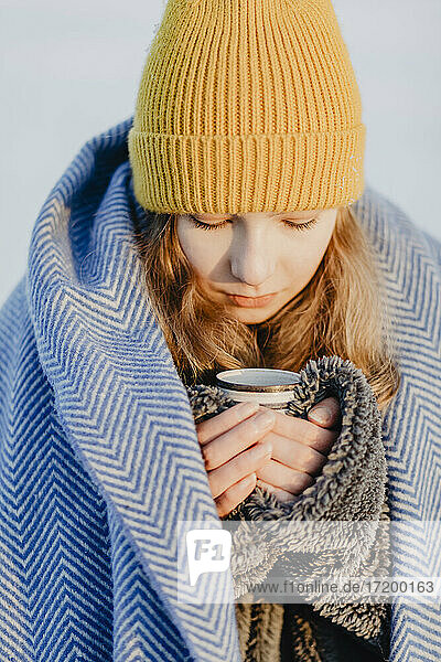 Porträt eines hübschen Mädchens im Teenageralter mit Strickmütze  das sich mit einer Wolldecke und einer Tasse heißem Tee wärmt
