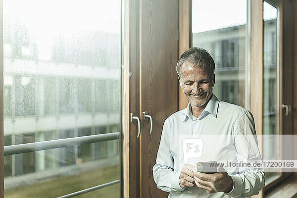 Smiling male entrepreneur using smart phone while standing at window in office