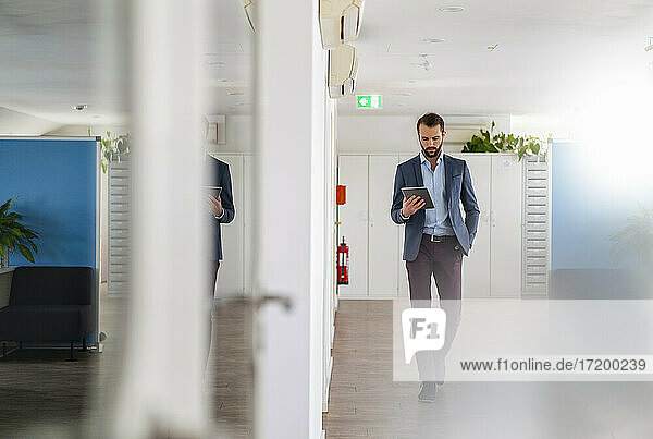 Geschäftsmann mit Hand in der Tasche  der beim Gehen im Büro ein digitales Tablet benutzt