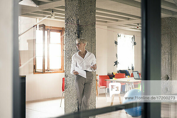 Geschäftsfrau durch Glaswand gesehen  während sie ein Dokument gegen eine Säule in ihrem Büro hält