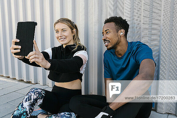 Young woman with man taking selfie through digital tablet while sitting on floor against wall
