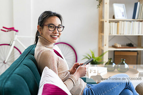 Junge Frau mit Brille sitzt mit Handy zu Hause