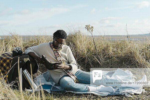 Young man reading book while sitting on blanket at beach against cloudy sky