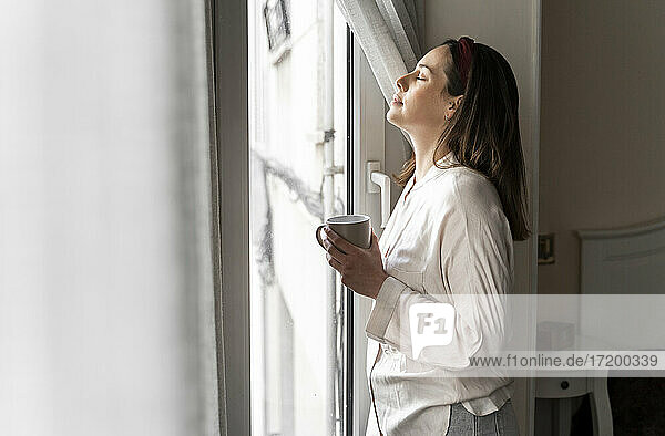 Entspannte junge Frau mit geschlossenen Augen  die eine Kaffeetasse hält  während sie am Fenster steht