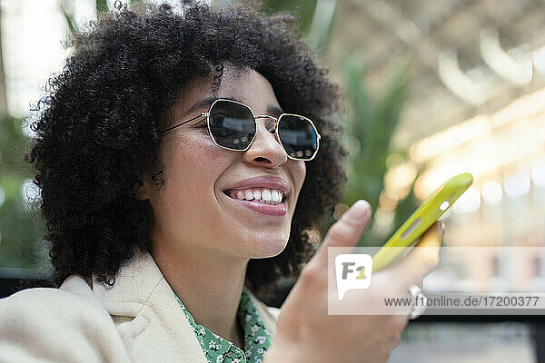 Lächelnde junge Frau mit Sonnenbrille und Smartphone in einem Café