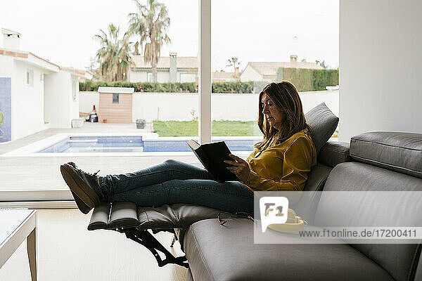 Entspannte Frau liest ein Buch  während sie zu Hause auf einem Lehnstuhl sitzt