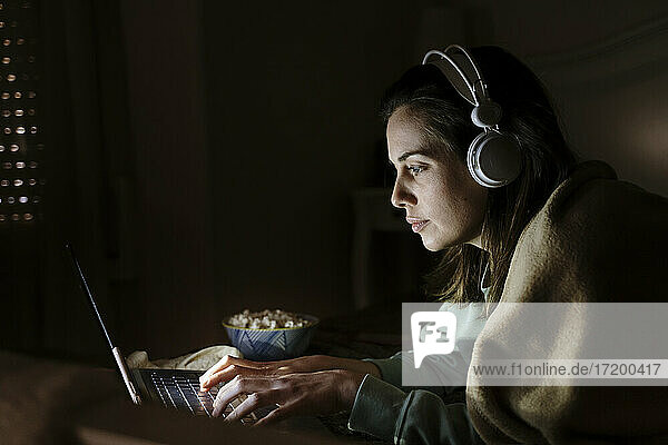 Frau mit Kopfhörern und Popcorn am Laptop  während sie zu Hause liegt