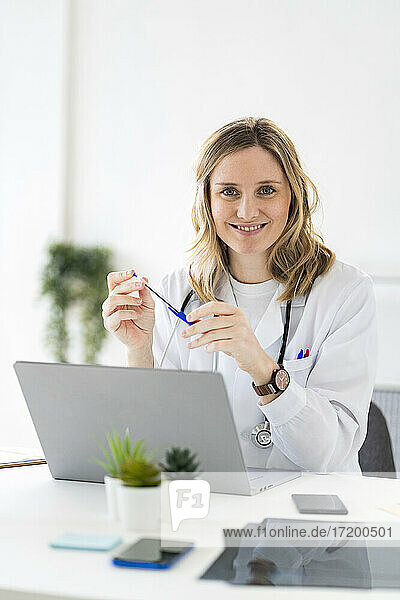 Lächelnde Ärztin sitzt mit Laptop am Schreibtisch in einer medizinischen Klinik