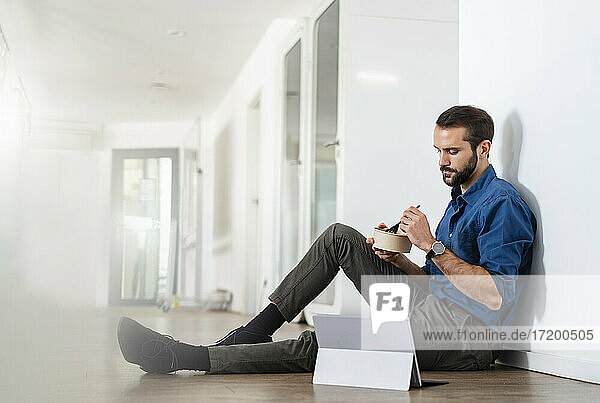 Geschäftsmann isst Essen  während er an einem digitalen Tablet auf dem Boden im Büro sitzt