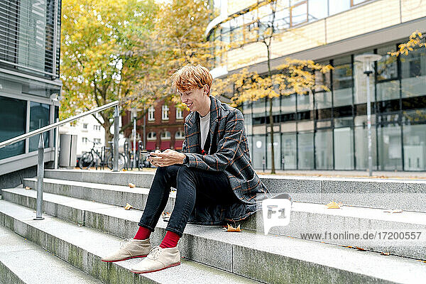 Smiling man using smart phone while sitting on steps against building