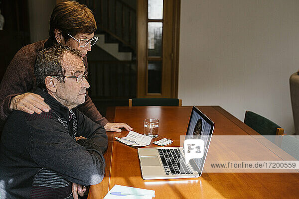 Eine Sozialarbeiterin steht neben einem älteren Mann  der sich mit einer Ärztin per Videoanruf über einen Laptop berät.