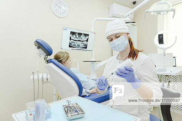 Zahnärztin mit Gesichtsschutzmaske bereitet Spritze für Injektion vor  im Hintergrund sitzt ein kleines Mädchen