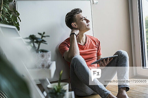 Nachdenklicher junger Mann mit digitalem Tablet  der wegschaut  während er im Wohnzimmer sitzt