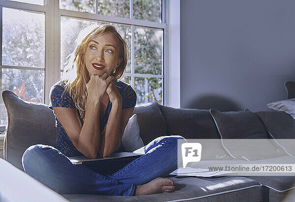 Nachdenkliche Frau schaut weg  während sie zu Hause mit dem Laptop auf dem Schoß sitzt