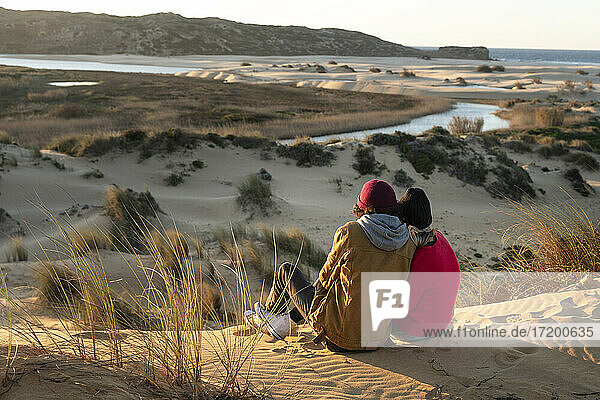 Paar mit Strickmütze sitzt bei Sonnenuntergang auf einer Sanddüne zusammen