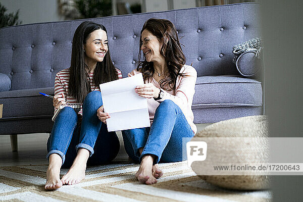 Mutter mit Dokument lächelt Tochter an  während sie im Wohnzimmer auf dem Boden sitzt