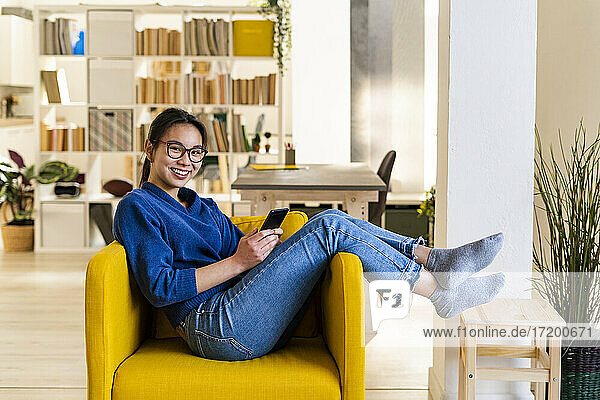 Lächelnde Frau mit Mobiltelefon auf einem Stuhl sitzend zu Hause