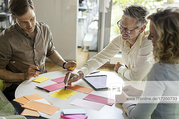 Älterer Geschäftsmann schreibt auf Papier  während er seinen Kollegen am Tisch im Büro die Geschäftsstrategie erläutert