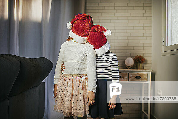 Sisters covering their faces with Santa hat while standing in living room